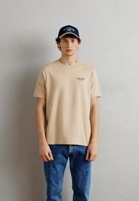Beige bomulds t-shirt med korte ærmer og rund halsudskæring, med lille sort tekst på venstre side. Båret med blå denim jeans.