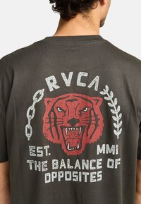 Donkergrijze t-shirt met een rode tijgerafbeelding, ketting en witte tekst die "RVCA EST. MMI THE BALANCE OF OPPOSITES" in een verouderde stijl weergeeft.