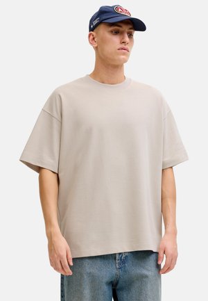 Young man wearing oversized beige t-shirt, light blue jeans, and navy blue cap with embroidered red and white logo, looking down.