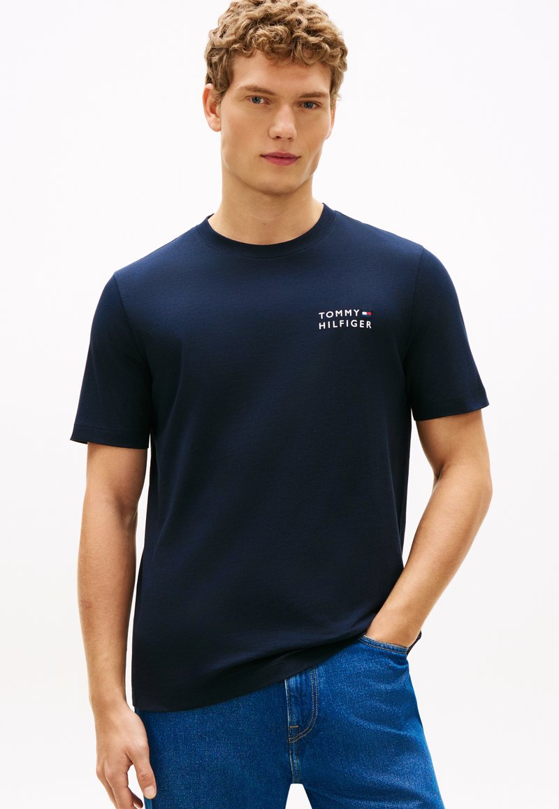 Νέος άνδρας με σγουρά μαλλιά φοράει μπλε σκούρο μπλουζάκι Tommy Hilfiger και μπλε τζιν, στέκεται με το ένα χέρι στην τσέπη.