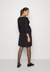 Vestido preto com mangas longas, corpete justo e saia rodada. Usado com meias pretas com bolinhas e sapatos planos, vista lateral do modelo.