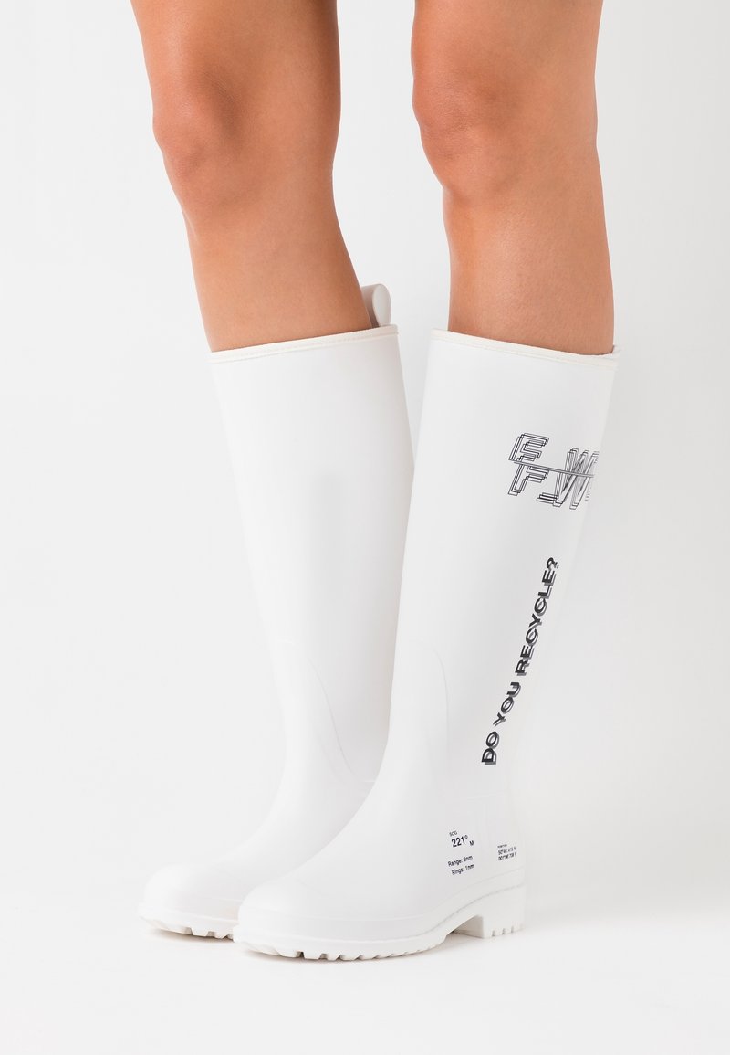 Bottes en caoutchouc blanches montantes jusqu'aux genoux portées sur des jambes nues, avec le texte noir "EST-CE QUE TU RECYCLÉS ?" sur le côté extérieur de la botte droite.