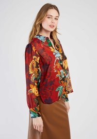 Rote Bluse mit floralem Muster und langen, gepufften Ärmeln, die grüne, gelbe und blaue Muster aufweist. Mit Kragen und Knopfdetails sowie einer lockeren Passform.