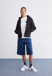 Haut à capuche noir zippé avec poches avant, t-shirt blanc avec un logo circulaire, shorts en denim bleu, chaussettes noires à rayures, baskets blanches.