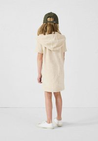 Vestido beige con capucha hecho de una tela suave, con mangas cortas y un corte relajado, combinado con una gorra verde y zapatillas blancas.