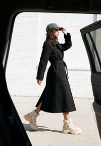 Femme en trench-coat noir, casquette grise et bottes beiges marchant devant un mur blanc en briques, vue à travers une portière de voiture ouverte.
