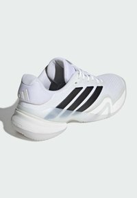 Biela športová obuv s čiernymi pruhmi, sieťovaným zvrškom, polstrovaným golierom a priehľadnou podrážkou. Má štruktúrovaný dizajn a tlmenú medzipodrážku.