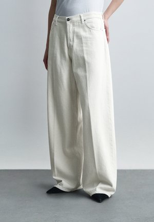 Kobieta w wysokich, szerokich, kremowobiałych dżinsach z czarnymi, spiczastymi butami, stojąca na tle jednolitego jasnoszarego tła.