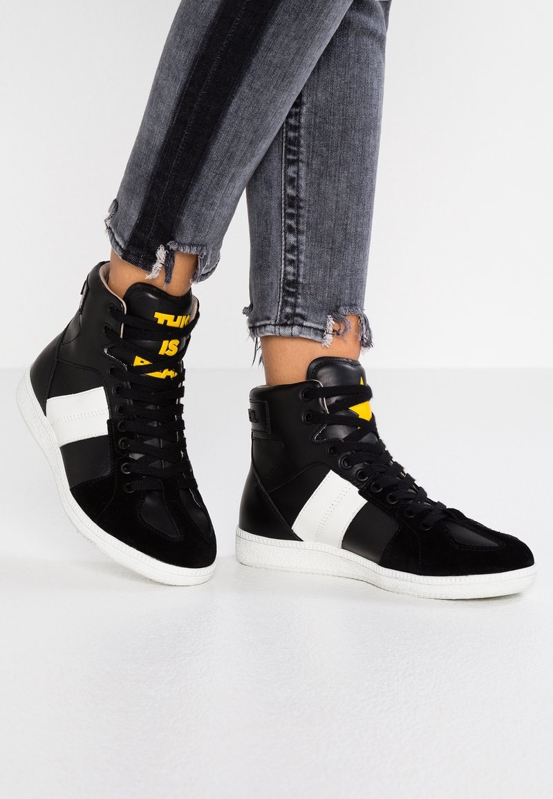 Vysoké tenisky vyrobené z černé kůže a semiše, s bílými akcenty a žlutým textem. Je vidět texturovaná podešev a potrhané džíny.