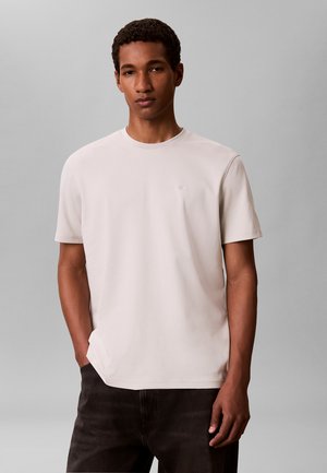 Ung mand iført en lysebeige t-shirt med korte ærmer og mørke bukser, stående foran en ensfarvet grå baggrund.