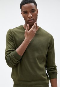 Sweter dzianinowy w oliwkowym kolorze z okrągłym dekoltem i prążkowanymi mankietami. Tkanina wydaje się miękka, ma gładką teksturę i dopasowany krój.