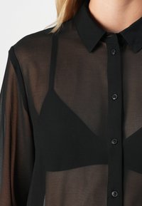 Gjennomsiktig svart skjorte med knapper og spiss krage, som avslører en svart cami som er brukt under.