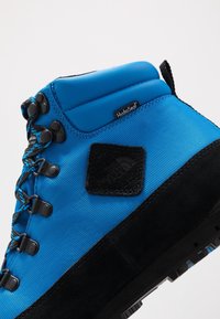 Modrá voděodolná turistická bota s texturovaným látkovým svrškem, černými semišovými detaily, kulatými tkaničkami a gumovou podrážkou s robustním vzorkem.