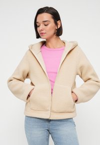 Μπεζ φλις μπουφάν με κουκούλα, διαθέτει φερμουάρ μπροστά και δύο μπροστινές τσέπες. Φοριέται πάνω από ένα ροζ μπλουζάκι και συνδυάζεται με ανοιχτόχρωμο μπλε τζιν.