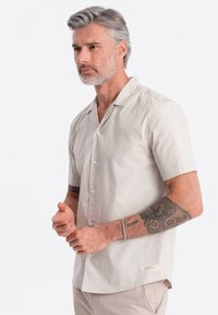 Kurzärmliges, kurzärmeliges Hemd aus leichtem beigen Stoff; glatte Textur mit Knopfverschluss und einem subtilen Markenetikett am Saum.