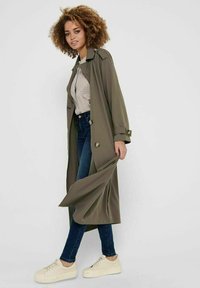 Olive green trench coat z długimi rękawami i guzikami, zestawiony z jasną koszulą w kratę, ciemnoniebieskimi dżinsami i kremowymi trampkami.