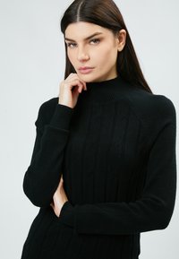 Maglione nero in maglia con colletto alto a costine, caratterizzato da motivi a treccia in rilievo e maniche lunghe. Texture morbida e design aderente.