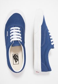 Blaue Wildledersneaker mit weißen Gummisohlen, ausgestattet mit weißen Schnürsenkeln und Nähdetails. Logo auf der Innensohle sichtbar.