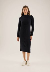 Eine Frau trägt ein langes, marineblaues Strickkleid mit goldenen Knöpfen an der Schulter und weißen Sneakern. Sie steht vor einem schlichten Hintergrund.