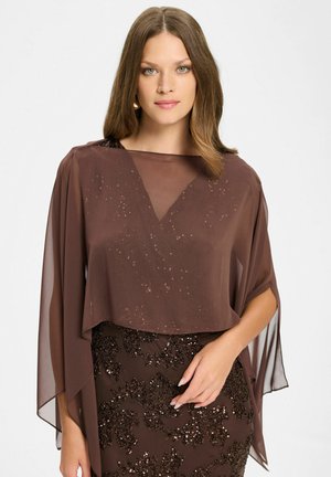 Kobieta z jasnobrązowymi włosami, ubrana w brązową, prześwitującą bluzkę nałożoną na sukienkę z cekinami w kwiatowy wzór, pozuje z jedną ręką na biodrze na białym tle.