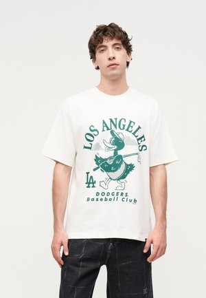 Junger Mann trägt ein weißes Los Angeles Dodgers T-Shirt mit einer Cartoon-Ente, die einen Golfschläger hält, kombiniert mit schwarzen Hosen.
