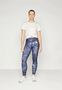 Tamno plave tajice s uzorkom i visokim strukom, izrađene od elastičnog materijala, u kombinaciji s svijetlosmešavom majicom s čvorom i bijelim sportskim cipelama.