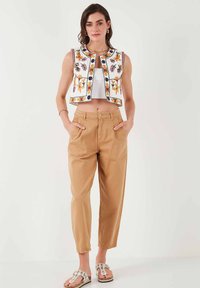 White embroidered vest with floral patterns, paired with tan tapered pants and embellished sandals. Smooth textures throughout.