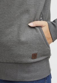 Felpa grigia realizzata in tessuto morbido, con una tasca testurizzata, una piccola etichetta in pelle e polsini a costine. Dettaglio di cucitura visibile sui lati.