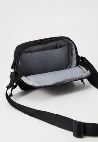 Geantă crossbody neagră cu închidere cu fermoar, având o căptușeală interioară gri, curea ajustabilă și un compartiment spațios fără buzunare externe.