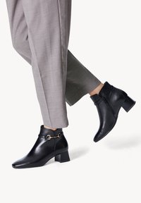 Black leather ankle boots with a pointed toe, side zipper, and a decorative buckle. Worn with gray tailored pants, showcasing a sleek design.