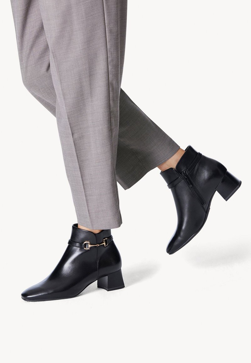 Black leather ankle boots with a pointed toe, side zipper, and a decorative buckle. Worn with gray tailored pants, showcasing a sleek design.