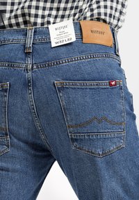 Niebieskie dżinsy denimowe o średnim stanie, slim fit. Posiadają dwie tylne kieszenie z kontrastującym żółtym przeszyciem i skórzaną metką marki. Widoczne metki.