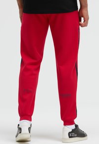 Rote Sweatpants mit dunklem Seitenpanel, elastischen Bündchen und Aufdruck. Kombiniert mit schwarz-weißen Sneakers, die eine glatte Textur aufweisen.