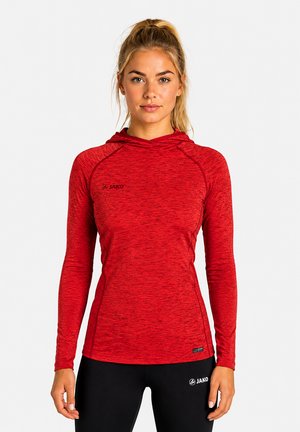 Donna con capelli biondi raccolti in una coda di cavallo, che indossa un top sportivo rosso aderente a maniche lunghe con cappuccio e pantaloni neri, in piedi davanti a uno sfondo semplice.