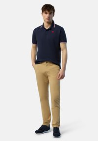 Polo shirt blu navy con colletto a righe rosse e bianche, pantaloni chino beige e sneakers nere. La maglietta presenta un piccolo logo sul petto.