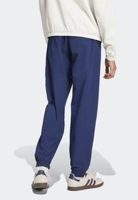 Námořnické modré tepláky z lehkého materiálu mají elastický pás a manžety. Modelka má na sobě bílé tenisky s barevnými pruhy.