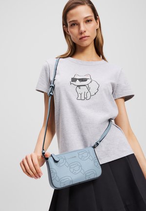 Borsa crossbody azzurro chiaro con motivo impresso e rifiniture nere. Pelle liscia con tracolla regolabile. Modello che indossa una maglietta grigia con grafica di un gatto.