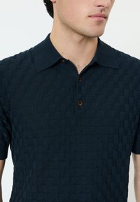 Moški v temno modri kratkoročni polo majici z reliefnim karo vzorcem in tremi rjavimi gumbi, prikazan od vratu do sredine trupa.