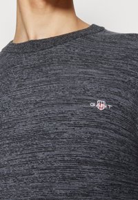 Gros plan d'une personne portant un pull en maille gris foncé avec un petit logo GANT et un emblème en forme de bouclier sur la poitrine.
