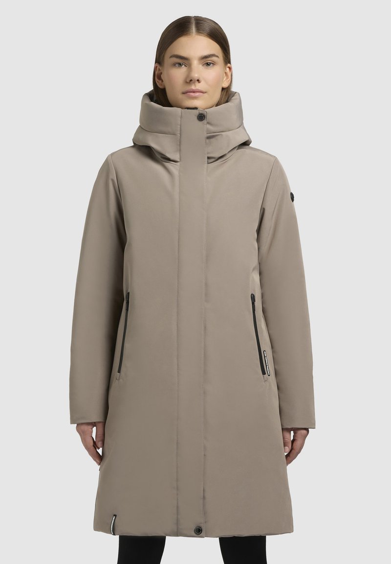 Beige, lange waterdichte jas met een hoge kraag, twee zijzakken met rits en een knoopsluiting aan de onderkant. Gladde textuur, minimalistisch ontwerp.