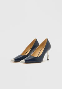 Marineblå skinnpumps med høye hæler, spiss tå og metallisk sølvhette. Har et elegant design og slank metallhæl.