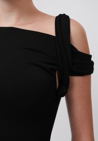 Svart topp med en glatt design, avtakbare skuldre og vridd tekstur ved skuldrene, laget av mykt og elastisk materiale.