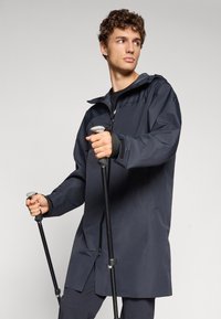 Mörkgrå vattentät jacka med huva, dragkedjestängning och justerbara ärmslut. Bäraren håller vandringsstavar, vilket visar på en funktionell design.