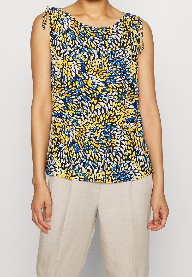 Haut sans manches avec un motif abstrait de feuilles jaune, bleu, blanc et noir porté avec un pantalon beige sur une personne debout.