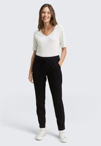 Witte V-hals top gecombineerd met zwarte broek met koord. De broek heeft zijzakken, een relaxte pasvorm en een gladde textuur. Sneakers in wit met zwarte accenten.