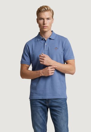 Giovane uomo con capelli biondi che indossa una polo blu a maniche corte e jeans blu, si aggiusta il polso sinistro con la mano destra.