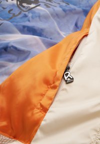 Farverig jakke med en glat orange kant, en sølvfarvet lynlås og et blåt mønstret indre med en subtil glans.