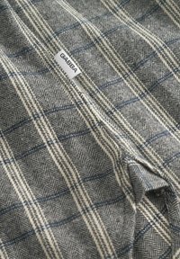 Țesătură din amestec de lână în model houndstooth gri și crem, cu linii de plasă albastre. O etichetă albă mică cu "GABBA" este vizibilă.