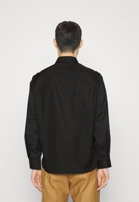 Camicia nera a maniche lunghe con colletto, texture liscia, polsini con bottoni e taglio dritto; indossata con pantaloni beige. Vista posteriore.