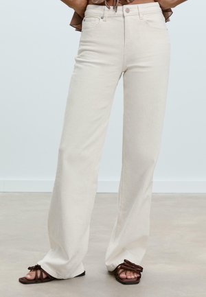 Jeans évasés crémeux taille haute avec une finition texturée, comportant des poches avant et une fermeture à bouton. Associés à des sandales à lanières brunes.